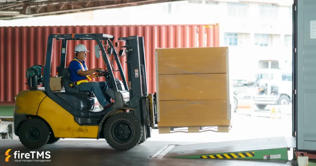 Operator kierujący wózkiem widłowym z ładunkiem, wjeżdżający po rampie magazynowej na pakę zestawu transportowego w jasny dzień